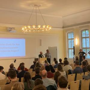 Zwei Personen die einen Vortrag gehalten haben an einem Pult, hinter ihnen eine Leinwand, mit einer Präsentation, vor Ihnen ein Publikum. Eine Person aus dem Publikum stellt eine Frage an die beiden Redner am Pult.