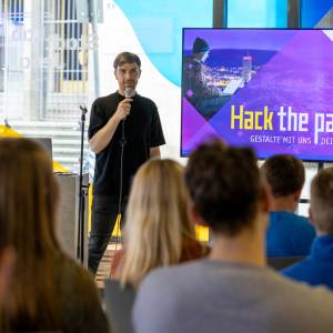 Moderator spricht vor Publikum mit einer Präsentation zum Hackathon "Hack the paradise!"