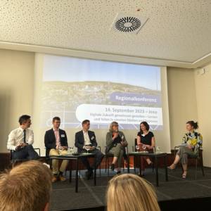 Podium mit sechs Personen vor einer Leinwand, vor ihnen das Publikum