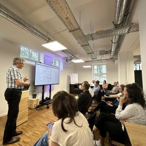 Martin Berger, Teilprojektleiter Handlungsfeld Digitale Infrastruktur und Datenpolitik, steht mit einem Bildschirm vor einem sitzendem Punblikum