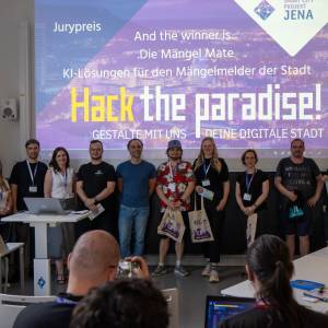 Vor einer Leinwand mit der Schrift "Hack the paradise! Gestalte mit uns deine digitale Stadt" stehen mehrere Menschen in einer Reihe mit Smart City Beuteln in der Hand vor einem Publikum
