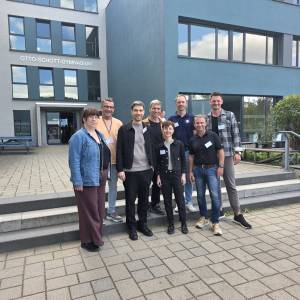 Gruppe von Menschen steht vor einer Schule und lächelt in die Kamera.