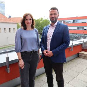 Mann und Frau stehen auf einer Dachterrasse mit Jena im Hintergrund und lächeln in die Kamera.