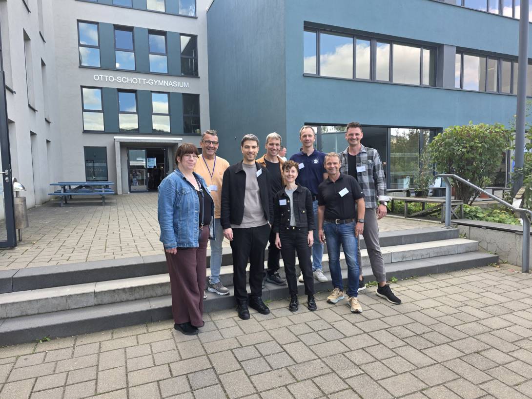 Gruppe von Menschen steht vor einer Schule und lächelt in die Kamera.