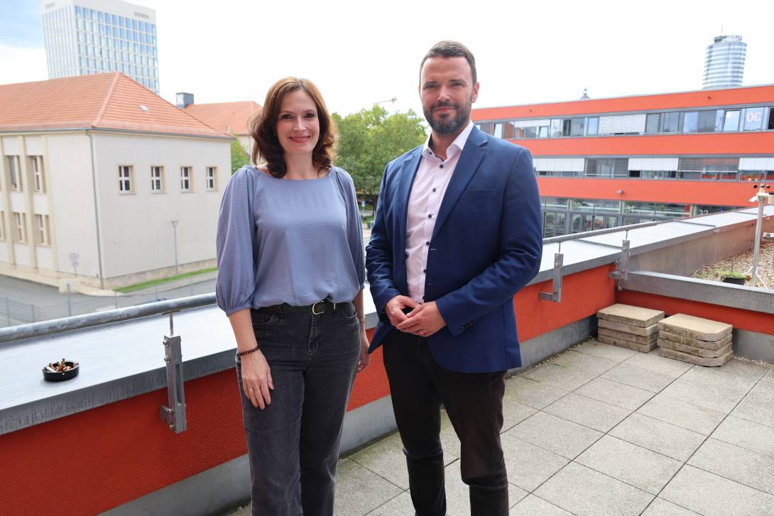 Mann und Frau stehen auf einer Dachterrasse mit Jena im Hintergrund und lächeln in die Kamera.